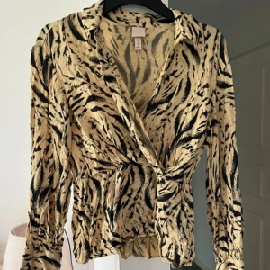 Beige blus med zebra-mönster från H&M - Säljer en beige blus från H&M med svart zebramönster. Blusen har omlott-design med knyt i midjan, långa ärmar med lätt puff och krage. Materialet är mjukt och lätt, perfekt för dig som gillar djurmönstrat och vill sticka ut.