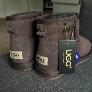BRUNA UGGS - Säljer ett par klassiska UGG boots i mörkbrun mocka med rund tå och platt sula. Skorna är fodrade med mjuk päls och har vattenresistent yta. Perfekta för kalla dagar och har UGG-logga baktill. HELT OANVÄNDA 