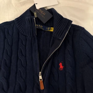 Marinblå stickad tröja Polo Ralph Lauren - Kabelstickad marinblå tröja från Polo Ralph Lauren med halv dragkedja och ribbad krage. Klassisk röd logga broderad på bröstet. Tröjan har långa ärmar och är tillverkad i mjuk ull, perfekt för kyliga dagar.