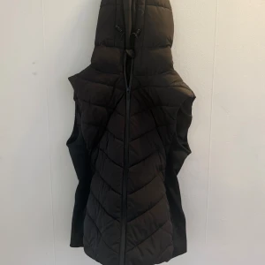 Svart quiltad dunväst från Zara - Svart lång dunväst från Zara med huva och dragkedja framtill. Västen är quiltad i ett snyggt mönster och har slitsar i sidorna för extra rörelsefrihet. säljer då det inte är stilen ja går för! skriv för mer bild!!