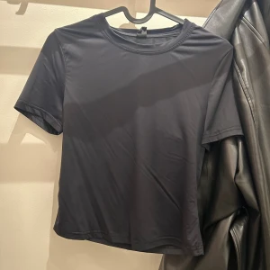 Svart croppad t-shirt i polyester - En stilren svart t-shirt med korta ärmar och rund hals. Modellen är croppad och har en slät, mjuk yta i polyester som ger en modern vibe. Perfekt att matcha med jeans eller skinnjacka för en enkel och cool look.