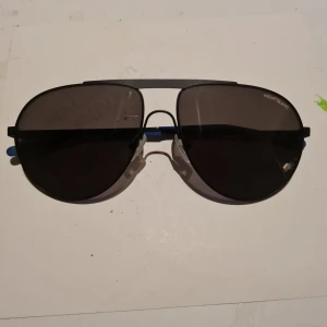 Svarta aviator-solglasögon Montblanc - Snygga svarta aviator-solglasögon från Montblanc, modell MB0119S. De har tunna metallbågar, mörka linser och blå detaljer på skalmarna. Klassisk och stilren design med Montblanc-logga på sidan. Perfekta för dig som vill ha en tidlös look.