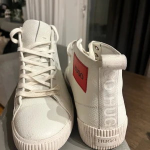 Vita high-top sneakers från Hugo - Snygga vita high-top sneakers från Hugo med röd logotyp på sidan. Skorna har snörning, rund tå och är tillverkade i präglat skinn med detaljer i textil vid sulan. Stilren design med ribbad sula och diskret Hugo-tryck på hälen.