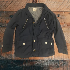 Mörkblå vindjacka från AIS denim laundry - Snygg mörkblå vindjacka från AJS med två bröstfickor med dragkedja och två stora framfickor med knappar. Saknas däremot en knapp. Kom gärna med bud och frågor om du har några.