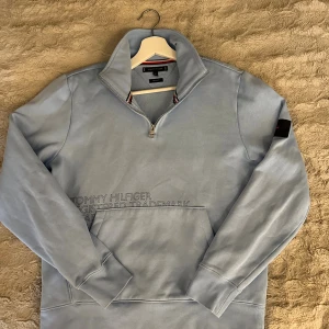 Ljusblå half zip från Tommy Hilfiger - Ljusblå sweatshirt från Tommy Hilfiger med half zip och hög krage. Regular fit med broderad logga på ärmen och klassisk magficka. Mjuk bomullskänsla och snygga detaljer i rött, vitt och blått på insidan av kragen.