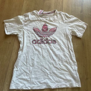 Vit Adidas t-shirt med rosa mönster - Snygg vit t-shirt från Adidas med det klassiska treklöver-loggan framtill, dekorerad med rosa och lila grafiskt mönster. Rund hals med matchande mönstrad kant. Perfekt för dig som gillar sportig streetstyle och vill sticka ut lite extra.