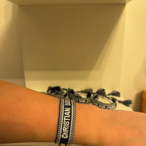 Svart Christian Dior armband - Snyggt svart armband från Christian Dior med vit broderad text och dekorativa tofsar i ändarna. Armbandet har en bred, vävd design och känns riktigt lyxigt och trendigt. Perfekt accessoar för att lyfta din outfit med en exklusiv touch.
