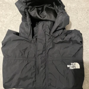 Svart vindjacka från The North Face - Svart vindjacka från The North Face med huva och meshfoder. Jackan har klassisk logga på bröstet och stängs med dragkedja och kardborre. Perfekt för blåsiga dagar och enkel att slänga på sig när vädret svänger.