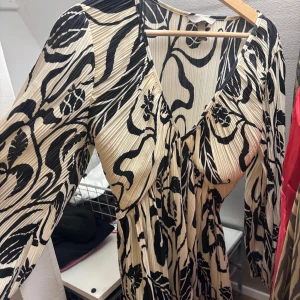 Plisserad svartvit klänning H&M S - Plisserad klänning från H&M i storlek S med svart abstrakt mönster på beige botten. Djup v-ringning, långa ärmar och lätt markerad midja. Jätte bra skick använd ett fåtal gånger
