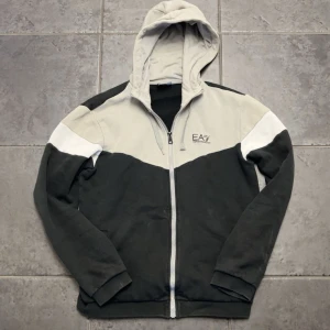 EA7 hoodie i svart, grå och vitt - Snygg hoodie från EA7 Emporio Armani i svart, grått och vitt. Tröjan har dragkedja framtill, huva och EA7-logga på bröstet. Tillverkad i mjuk bomullsmix, perfekt för chill eller streetwear. Långärmad med ribbade muddar och cool färgblock-design.
