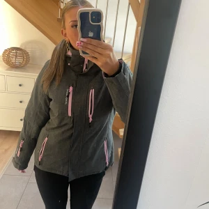 Grå vinterjacka Swedemount med rosa detaljer - Säljer en grå vinterjacka från Swedemount med rosa dragkedjor och detaljer. Jackan har huva, flera fickor med dragkedja och hög krage. Perfekt för kalla dagar och har en snygg, sportig look. Passformen är normal och jackan är långärmad.