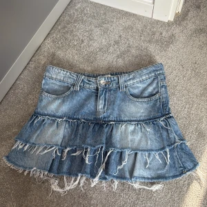 Blå jeanskjol med volanger från Gina Tricot - Säljer en blå jeanskjol från Gina Tricot med två lager volanger och fransad nederkant. Kjolen har klassiska jeansdetaljer som fickor och bälteshällor, och är gjord i bomull. Perfekt för dig som gillar en cool och avslappnad stil.