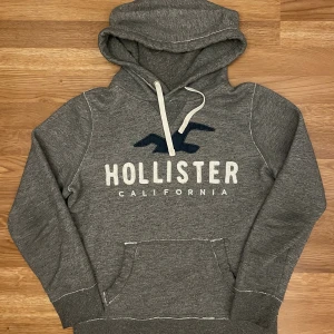Grå hoodie från Hollister XS - Mysig grå hoodie från Hollister med stor logga och broderad fågel framtill. Tröjan har huva med vit snörning och känguruficka. Perfekt för kyliga dagar och chill vibes. Tillverkad i mjukt material som känns skönt mot huden.