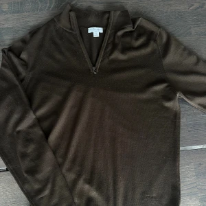 Brun merinoullströja från Sunspel - Snygg brun långärmad tröja från Sunspel i 100% merinoull. Tröjan har en stilren krage med half zip-dragkedja och en klassisk passform. Perfekt för dig som gillar en clean och tidlös look.