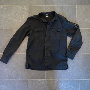 Svart overshirt från C.P. Company - Jätte snygg jacka från Cp company som är i ett bra skick. Jackan har inga större fläckar eller annat, men har några enstaka repor på badgen (se bild 4) Jackan är storlek L, MEN har passform som M (även därför jag säljer) Fråga!