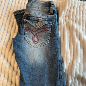 Blå bootcut jeans från Gina Tricot - Snygga blå jeans från Gina Tricot med bootcut passform, låg midja  och slitna detaljer på knäna. Jeansen har klassiska fem fickor och cool röd brodering på bakfickan. Materialet är jeans med ljus tvätt och kontrastsömmar.