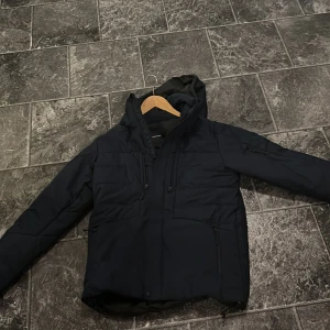 Svart skidjacka Peak Performance - Snygg svart skidjacka från Peak Performance med huva och flera dragkedjeförsedda fickor. Jackan har en stilren design med quiltade partier och justerbar dragsko vid huvan. Perfekt för kalla dagar när du vill hålla värmen och ändå se fräsch ut. Knappt använd, inköpt för 3800