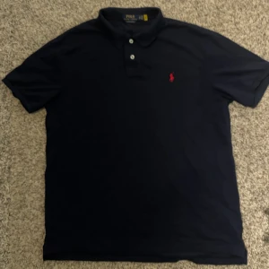 Mörkblå pikétröja Polo Ralph Lauren L - Klassisk mörkblå pikétröja från Polo Ralph Lauren i slim fit med två knappar och röd broderad logga på bröstet. Tröjan har korta ärmar och mjukt bomullsmaterial som känns skönt mot huden. Perfekt för en clean och stilren look. Aldrig Använd. Kontakt mig gärna för övriga frågor🙌 Passar mer L än XL
