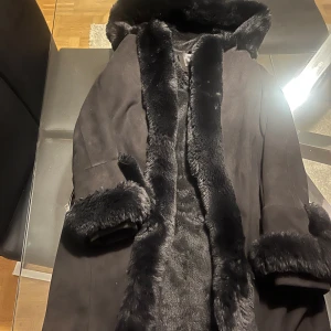 Svart pälsjacka från Garoff med huva - Snygg svart pälsjacka från Garoff med stor fluffig huva och breda pälskanter längs öppningen och ärmsluten. Jackan har en längre modell och ger en lyxig look. Saknar dock band till midjan men går lika bra med ett band från annan kappa man har hemma!