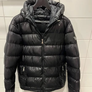 Svart pufferjacka Alessandro Zavetti - Snygg svart pufferjacka från Alessandro Zavetti med huva och dragkedja framtill. Jackan har två fickor med dragkedja och diskret logga på bröstet samt ärmen. Perfekt för kalla dagar och riktigt stilren look.