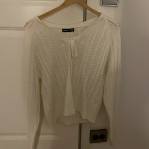 Vit stickad kofta med knyt från SHEIN - Supersöt vit stickad kofta från SHEIN med snyggt knyt framtill. Koftan har ett flätat mönster och långa ärmar, perfekt att slänga över en topp. Den är croppad och har en mjuk känsla som gör den skön att bära både inne och ute.
