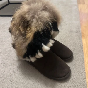 Bruna boots med fluffig päls - Unika bruna boots med rund tå och platt sula, dekorerade med tjock, flerfärgad päls i beige, svart och vit. Perfekta för dig som vill sticka ut och hålla dig varm under vintern. Materialet är mocka och päls, och de har en riktigt cozy vibe. Finns liten fläck från snö