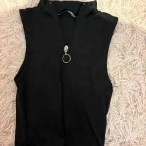 Svart ribbad ärmlös topp från lager 157 - Säljer en svart ribbad ärmlös topp från lager 157 med hög krage och cool dragkedja framtill med ringdetalj. Toppen har en slim passform och är perfekt för att skapa en trendig look. Supersnygg att styla med jeans eller kjol! 