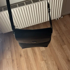 Svart axelväska från Emporio Armani - Snygg svart axelväska från Emporio Armani i skinn med präglat mönster och logga framtill. Väskan har justerbar axelrem, magnetlås och dragkedjefack på baksidan. Perfekt för dig som vill ha en stilren och praktisk väska med exklusiv känsla.