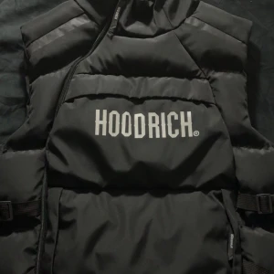 Svart dunväst från Hoodrich - Säljer en svart dunväst från Hoodrich i storlek M. Västen har en clean look med tydlig logga framtill, quiltad design och dragkedja. Materialet är syntet och polyester, perfekt för lager-på-lager-stil. Snygg och enkel att matcha till streetwear.