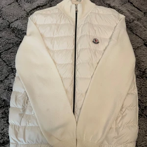 Vit Moncler dunjacka med stickade ärmar - Snygg vit dunjacka från Moncler med quiltad front och rygg samt stickade ärmar och krage. Jackan har dragkedja framtill och Moncler-logga på bröstet. Perfekt för kyliga dagar när du vill ha både stil och värme. Lite fläkt på jackan men syns knappt 