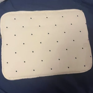 Vit prickig datorväska med dragkedja - Säljer en stilren vit datorväska med svarta prickar och dragkedja runt kanten. Väskan har en mjuk och vadderad känsla, perfekt för att skydda din laptop. Enkel och snygg design som passar till det mesta. Den är helt ny