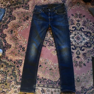 Mörkblå slim jeans från Nudie Jeans finn 28/32 snygg fade - Snygga mörkblå jeans från Nudie Jeans med skinny passform och klassisk femficksdesign. Jeansen har slitningar och kontrastsömmar, samt Nudie-logga på bakfickan och läderpatch i midjan. Perfekt för dig som gillar en smal siluett och coola detaljer. Midja 36cm längd 103cm (32)