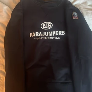Svart fleece-tröja från Parajumpers - Svart fleece-tröja från Parajumpers med stort vitt tryck på bröstet och broderad patch på ärmen. Klassisk rund hals och ribbade muddar. Perfekt för dig som gillar streetwear och vill ha något både snyggt och bekvämt.