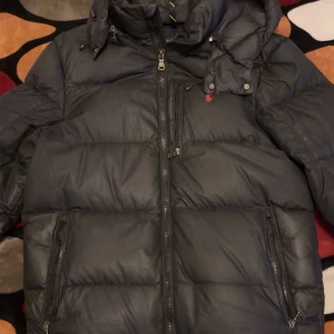 Svart dunjacka från Polo Ralph Lauren - Säljer en svart dunjacka från Polo Ralph Lauren med huva och dragkedjor. Jackan har en klassisk pufferstil, lång ärm och diskret logga på bröstet. Perfekt för kalla dagar och riktigt snygg till streetwear-looken. Har ett litet hål på ryggen som är sytt, och den är äkta