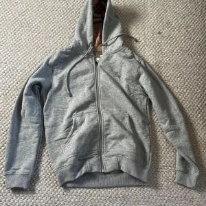 Grå hoodie från Burberry med dragkedja - Grå hoodie från Burberry med klassiskt rutigt foder i huvan och insidan. Tröjan har dragkedja, snörning i huvan och broderad logga på bröstet. Tillverkad i mjuk bomullsmix och har känguruficka framtill. Perfekt för en chill och stilren look.