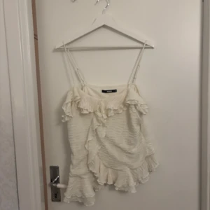 Vit volang topp med smala axelband - Supersöt vit topp från Bik Bok med smala axelband och volangdetaljer både upptill och nertill. har en lätt rynkad struktur och känns riktigt somrig och luftig. Perfekt för dig som gillar romantisk och feminin stil.