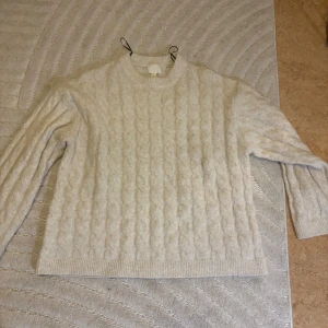 Beige kabelstickad tröja - Kabelstickad beige tröja med rund hals och långa ärmar. Tröjan har ett klassiskt flätmönster och ribbade muddar vid ärmslut och nederkant. Perfekt för kyliga dagar och enkel att matcha med jeans eller kjol. Använd fåtal gånger