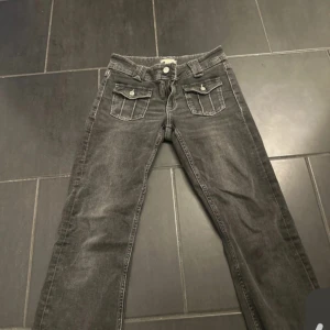 Grå jeans med fickdetaljer - Snygga grå jeans med två framfickor med lock och knappar. Jeansen har låg midja och raka ben, perfekt för en avslappnad stil. Materialet är klassisk denim och färgen är tvättad grå med lätt slitning.