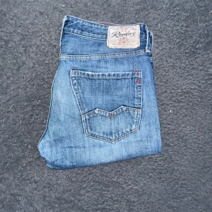 Replay Waitom jeans  - Säljer ett par klassiska blå jeans från Replay med raka ben och snyggt slitna detaljer. Jeansen har fem fickor, normal passform och tydliga Replay-loggor på bakfickorna och patchen. Perfekta för dig som gillar en tidlös och avslappnad look. Längd 99cm, midja 45cm. JAG SKICKAR INGA FLER BILDER NÄR JAG BÄR JEANSEN ! OBS som man ser på sista så har dem fått en liten reva för bak på ca 2 cm och ett pyttelitet hål i revan, i övrigt i mycket bra skick !