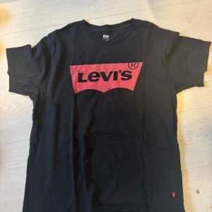 Svart Levi's t-shirt med röd logga - Svart t-shirt från Levi's i storlek L med klassisk röd logga framtill. T-shirten har rund hals och korta ärmar, tillverkad i mjuk bomull. Perfekt för dig som gillar streetwear och vill ha en enkel men snygg look.