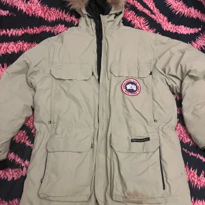Beige Canada Goose parkas med päls - Säljer en beige parkas från Canada Goose med ikonisk röd och vit patch på bröstet. Jackan har stor huva med päls, flera praktiska fickor och dragkedja framtill. Perfekt för kalla vinterdagar och har en klassisk, oversized look.