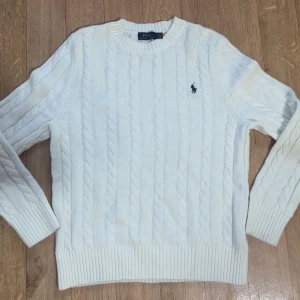 Vit kabelstickad tröja Polo Ralph Lauren - Vit kabelstickad tröja från Polo Ralph Lauren med klassisk rund halsringning och broderad marinblå logga på bröstet. Tröjan har ribbade muddar och är tillverkad i mjuk bomull. Perfekt för lager-på-lager och snygg till jeans eller kjol.