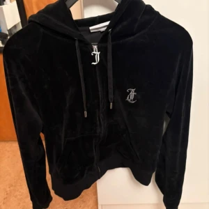 Svart velour hoodie från Juicy Couture - Svart hoodie i mjuk velour från Juicy Couture med glittrande Juicy-text på ryggen och JC-logga framtill. Tröjan har huva, dragkedja och snörning med metalländar. Perfekt för en chill och trendig look.