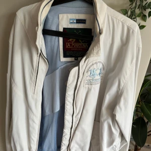 Vit vindjacka från La Martina XL - Säljer en stilren vit vindjacka från La Martina i storlek XL. Jackan har dragkedja framtill, ribbad nederkant och broderad logga på bröstet. Lätt och sportig design med meshfoder på insidan, perfekt för dig som gillar klassisk och clean look. ny pris= ca 5000