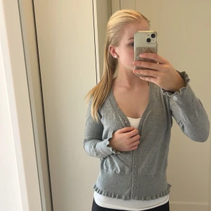 Grå kofta med volangkant från Lindex - Grå kofta från Lindex i mjukt material med v-ringning och knappar framtill. Långa ärmar och volangdetaljer vid ärmslut och nederkant ger en söt touch. Perfekt att slänga över ett linne eller t-shirt för en chill look.