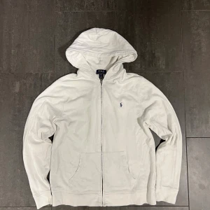 Ralph Lauren zip  - Vit hoodie från Polo Ralph Lauren i storlek XL. Klassisk design med dragkedja, huva och känguruficka. Broderad blå logga på bröstet. Tillverkad i mjuk bomull, perfekt för en chill och stilren look. Storlek barn xl sitter som S/M. Tveka inte att höra av er vid frågor och funderingar och kolla gärna in mina andra annonser så kan vi säkert fixa ett bra paketpris🤩🤩🤩