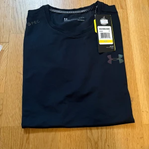 Svart Under Armour t-shirt  - Svart t-shirt från Under Armour med diskret logga på bröstet och UA-SPEC-tryck på axeln. Tillverkad i funktionsmaterial HeatGear som andas och passar perfekt för träning. Klassisk rund hals och korta ärmar. Snygg och sportig stil.