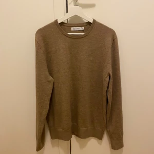 Beige Calvin Klein tröja i ullmix - Snygg beige tröja från Calvin Klein med diskret CK-brodyr på bröstet. Tröjan har rund halsringning och är tillverkad i en mjuk ull- och polyestermix som känns både lyxig och bekväm. Perfekt för lager-på-lager under höst och vinter.