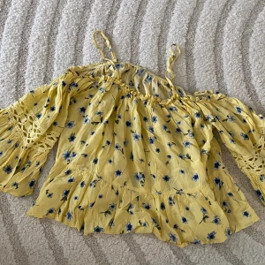 Gul blommig blus med spetsdetaljer - Supersöt gul blus från H&M med blått blommönster och volangkant. Blusen har långa ärmar med virkade spetsdetaljer och smala axelband. Perfekt för vår och sommar när du vill ha en romantisk och bohemisk vibe.