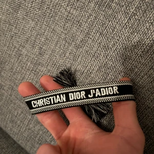 Christian Dior J'Adior armband svart - Snyggt svart och vitt armband från Christian Dior med texten 'J'Adior' broderad. Armbandet har fransar i ändarna och är perfekt för att ge din outfit en lyxig touch. Klassisk och trendig accessoar från Dior.